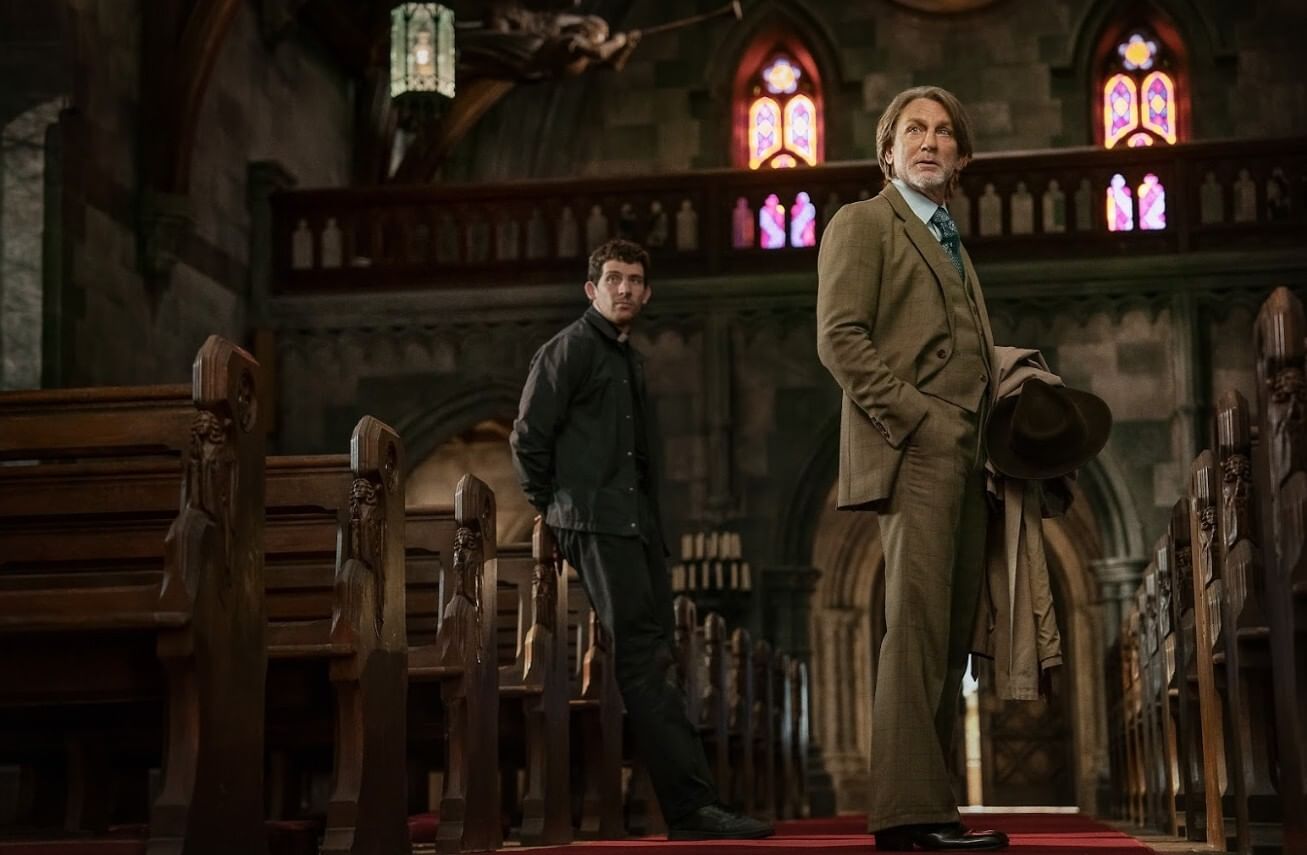 Craig, in a snazzy brown suit, stands in a small, ornate Catholic nave with wooden pews, stone arches, and stained glass.