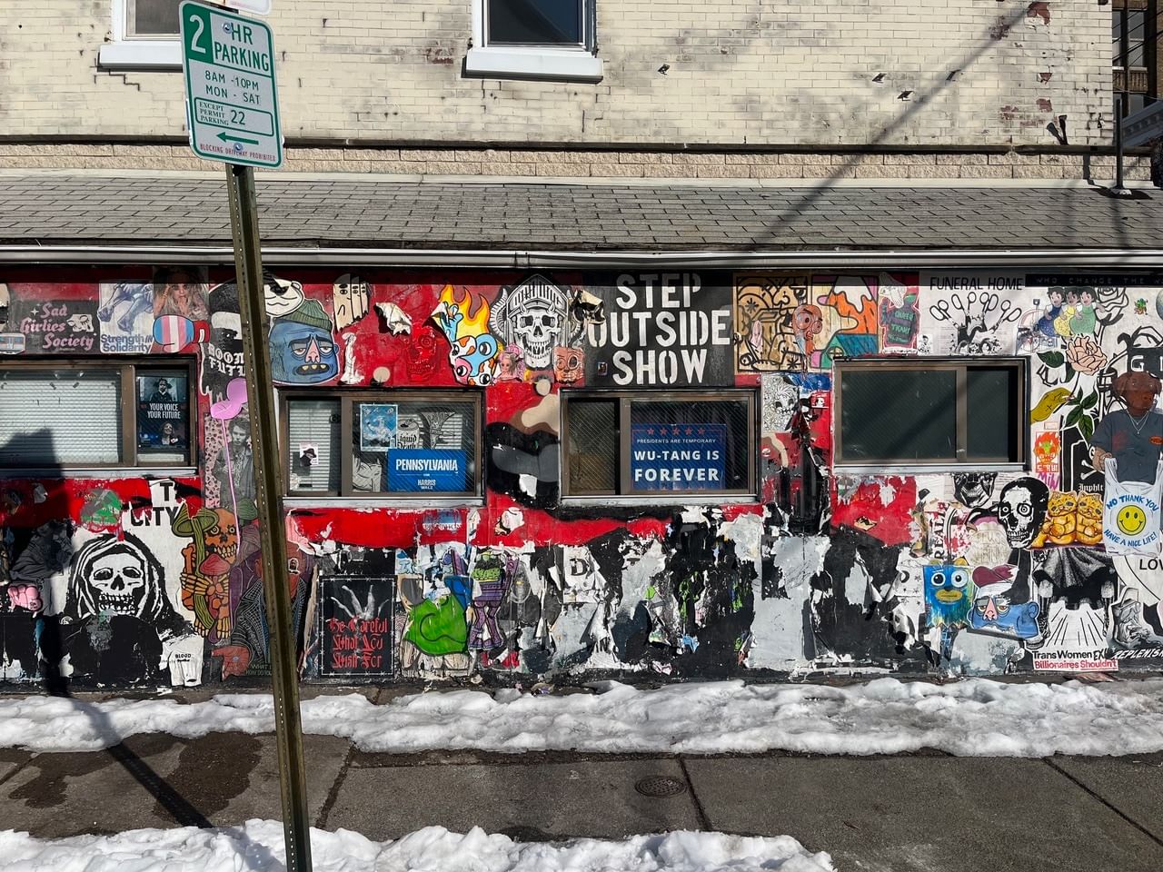 View from the sidewalk of a low city building chaotically covered with colorful stickers and decaying wheat-pastes.