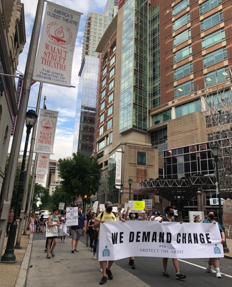 A large crowd of summertime protestors, holding a We Demand Change banner, walk on 9th Street past the Walnut Street Theatre.