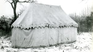 Black and white photo of a large tent in a snowy, wooded area, cords holding it up