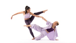 Two dancers, all white background, in mid-motion. One stands, holds the other dance up from the floor by one hand