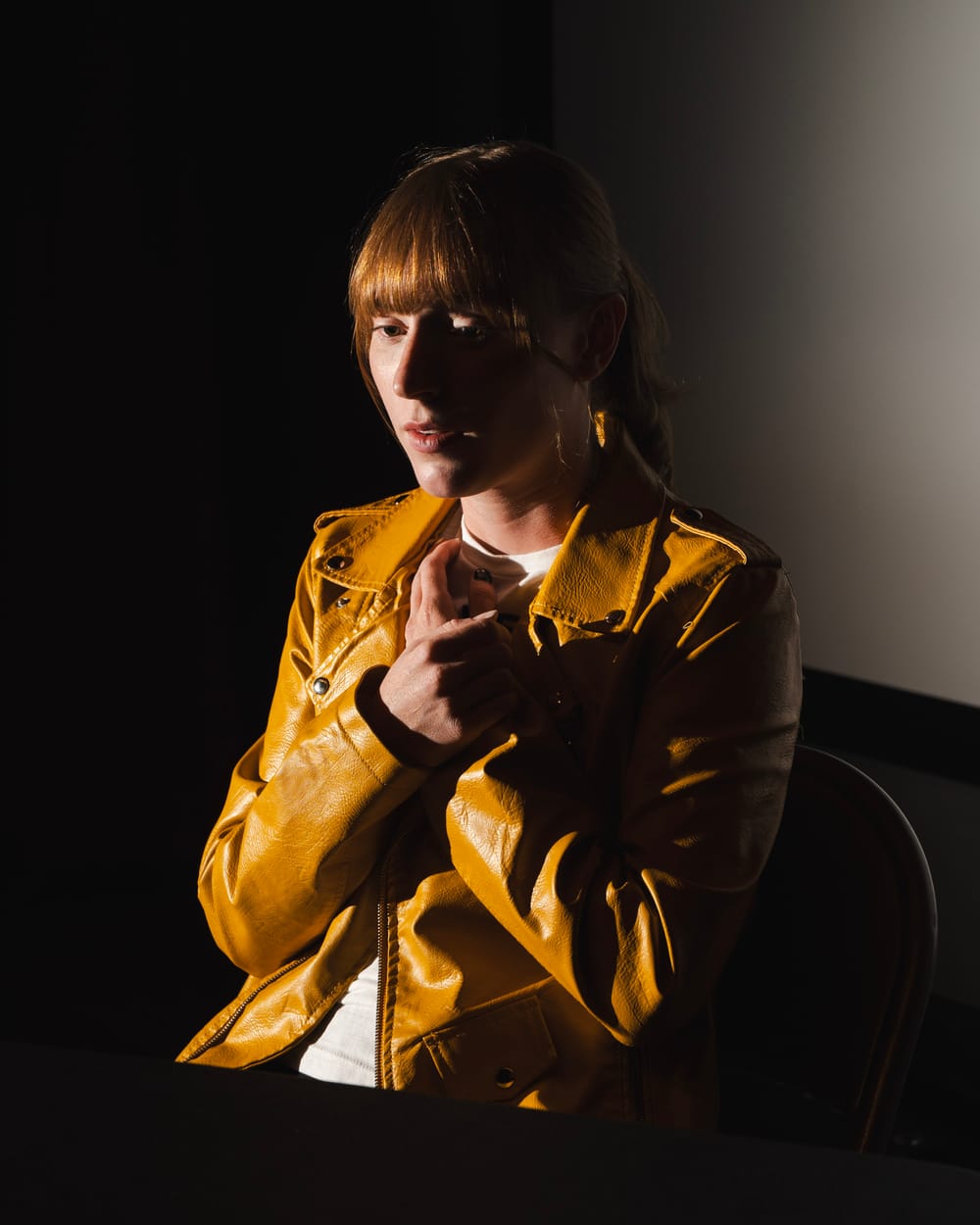 Webeloff, a white woman with auburn hair and yellow leather jacket, sits with hands clasped to her chest, a worried gesture