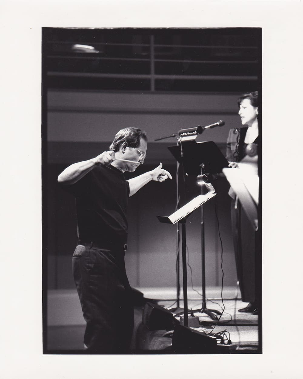 A black and white photo of Krzywicki, in all black, his arms and hands out as he conducts soprano Kidwell in front of him