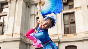 Ma, a Chinese woman in flowing blue & pink traditional garb poses smiling with a blue fan above her head outside City Hall.