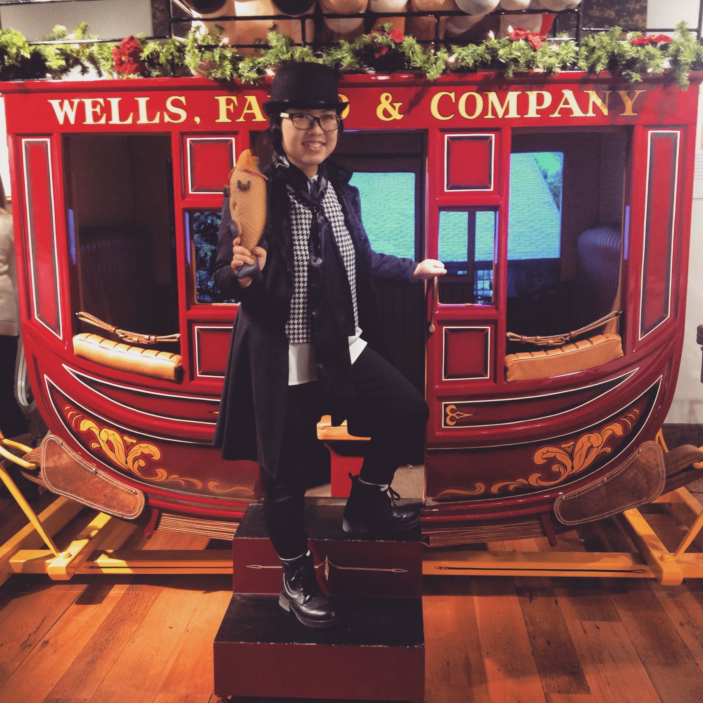 Bao, a Chinese woman in glasses, long coat, black boots, and black hat, poses smiling next to a red Wells Fargo carriage.