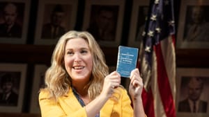 Actor Cassie Beck, a white woman, wears a yellow blazer and holds a pocket-sized US Constitution, in front of a US flag