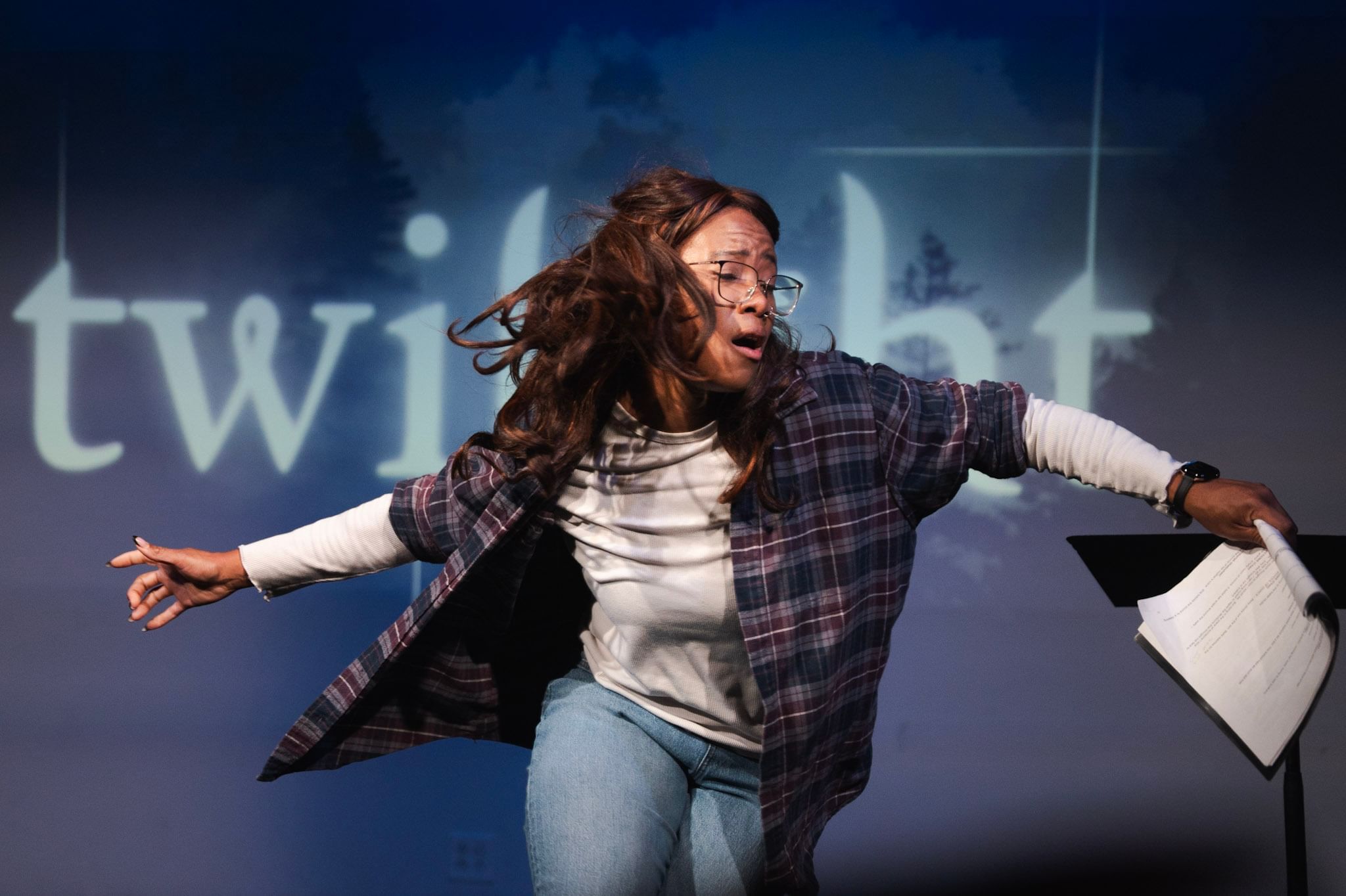 Sconiers, a Black woman with long brown hair flying around her and a flannel shirt, pivots dramatically, holding a script
