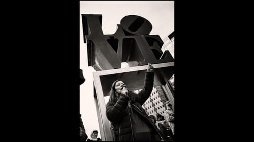 Black & white photo of a woman in a black winter jacket speaking with a fist raised under the LOVE statue.