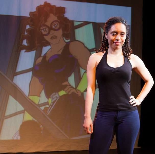 A Black woman stands in front of a projected image of a Black super hero woman wearing an eye mask and black outfit.
