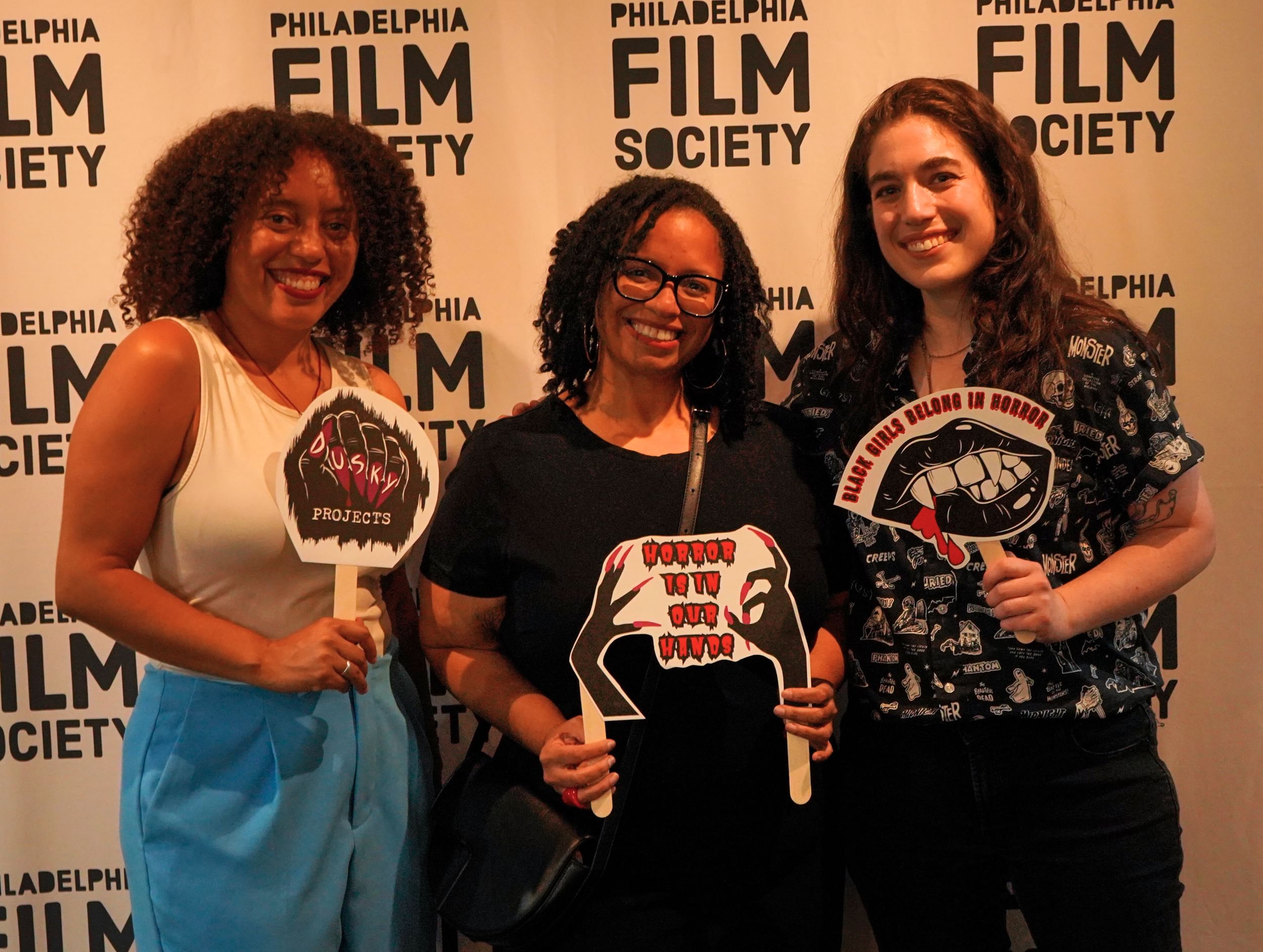 Two Black women and one white woman smile standing in front of a backdrop that says "Philadelphia Film Society."