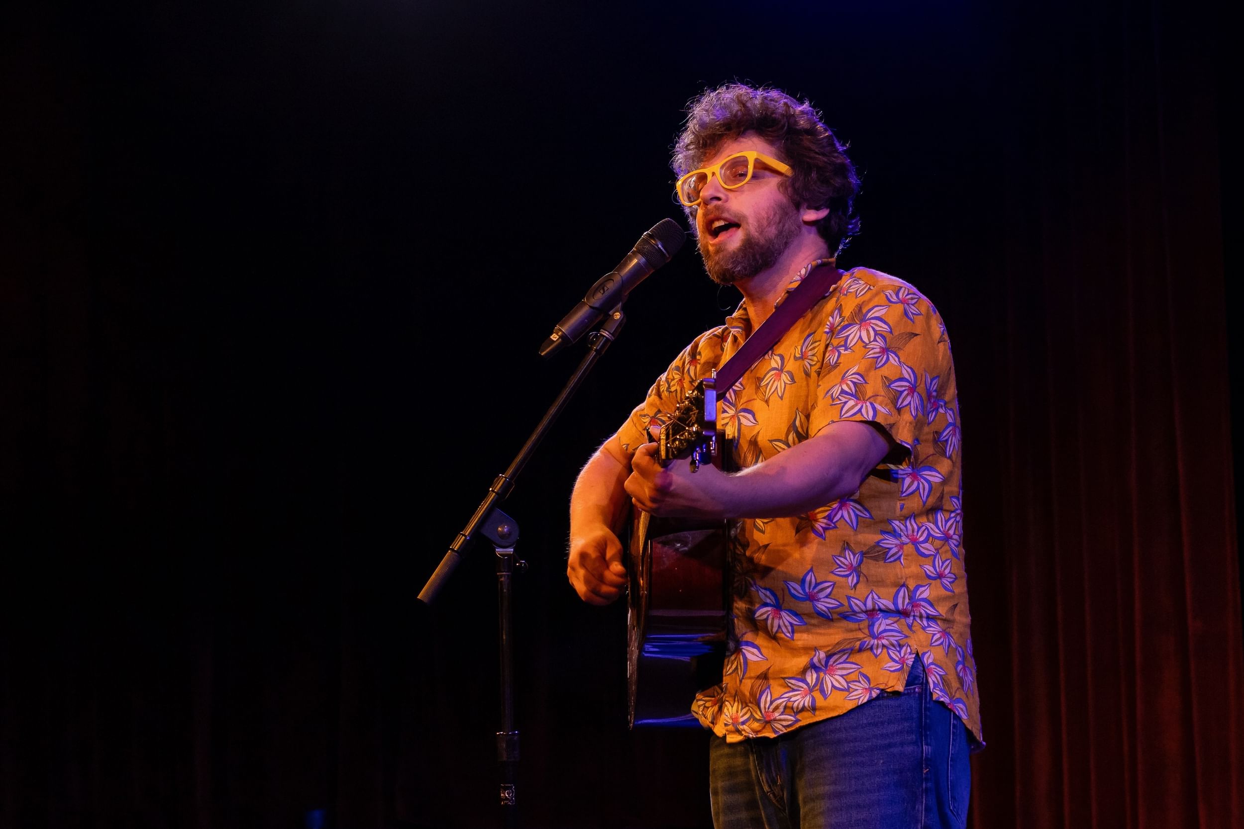 Osband, a bearded white man with yellow glasses and a colorful shirt, strums a guitar and sings into a mic.