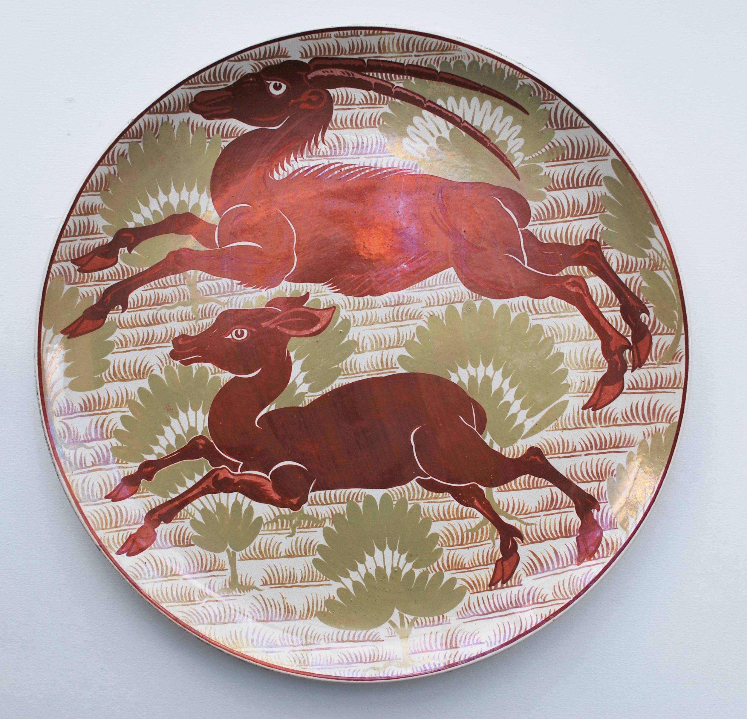 A lustrous ceramic plate painted with a gold foliage backdrop and leaping antelopes in profile, in shimmering burgundy.