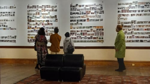 Gallery view showing large pieces of paper holding dozens of photos mounted in rows; four people look at them.