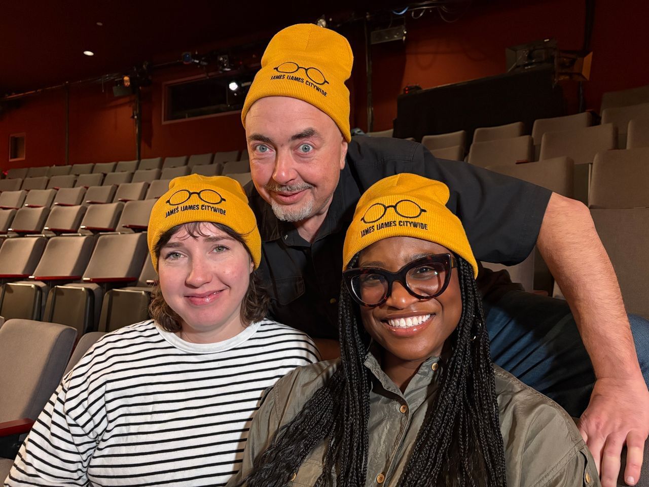 Urnov, Green, and Robinson smile together in the Wilma’s empty house, rows of seats behind. They wear yellow Ijames beanies