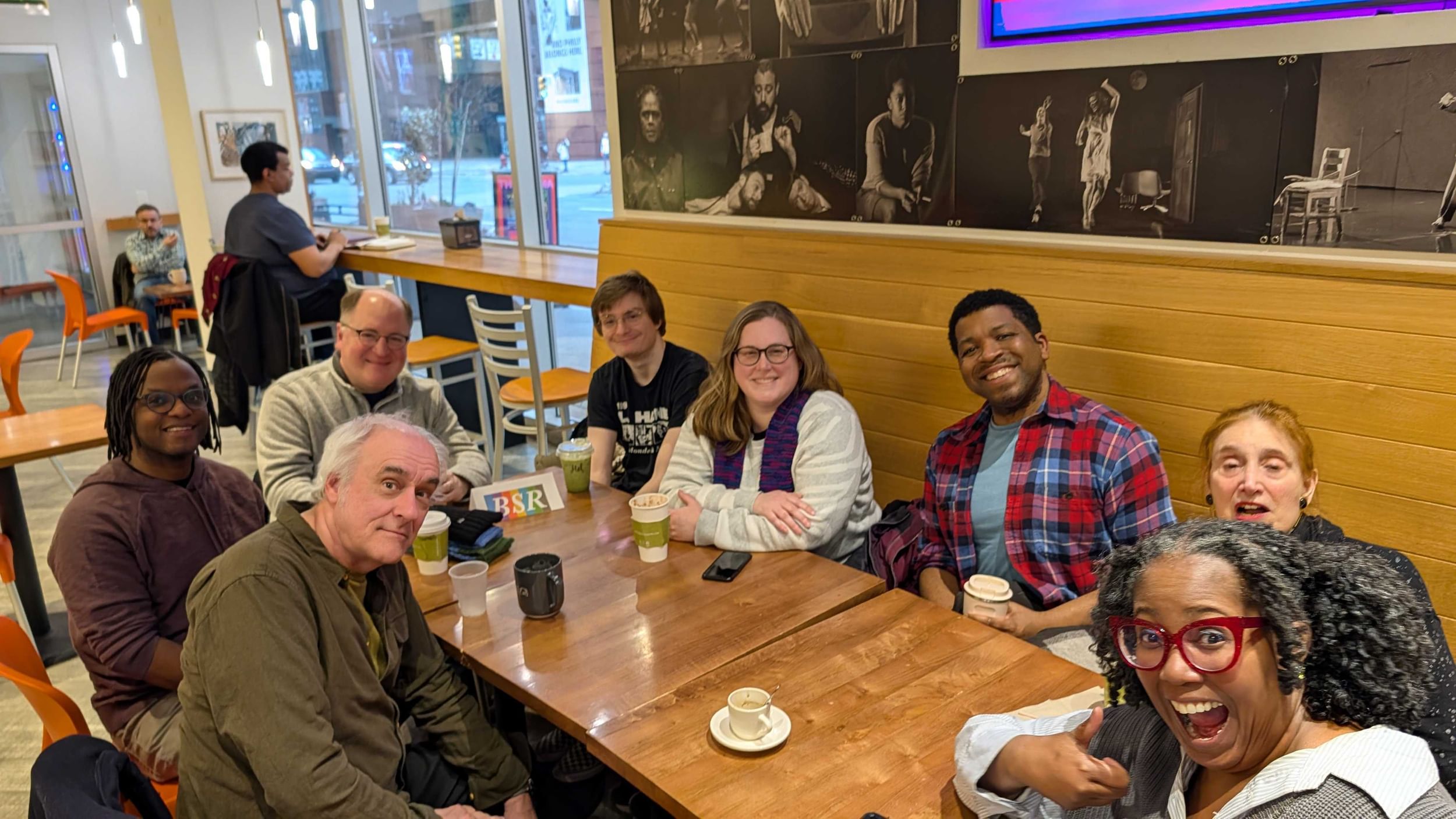 Diverse group of 8 people in casual clothes around a cafe banquette table, wearing casual clothes and smiling.