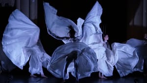 Three dancers in flowing white robes that expand like wings. Dimly lit, the robes are visible, the dancers are almost not