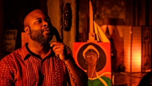 In dramatic red lighting, Davis listens to the phone he holds to his ear. A painting of a Black woman is behind him.