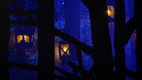Closeup on a layered shadowbox design with glowing lanterns against foreboding blue and black trees.