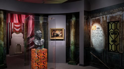 A bust of Washington in a vitrine on a Kente cloth style patterned pedestal around other historic depictions & artifacts.