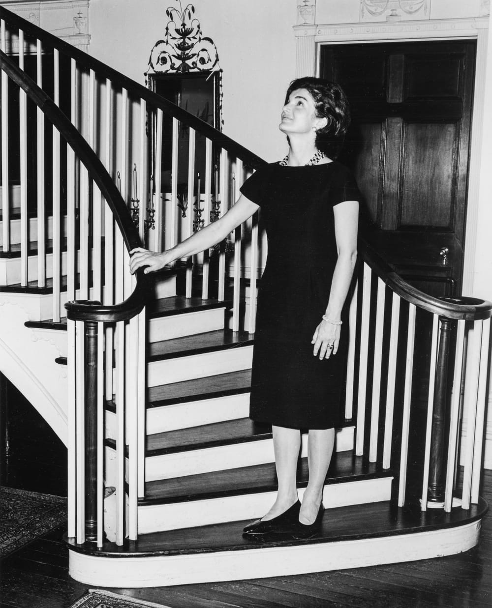 A black & white photo of Kennedy, standing at the bottom of Winterthur’s grand curving stairway, looking admiringly upward