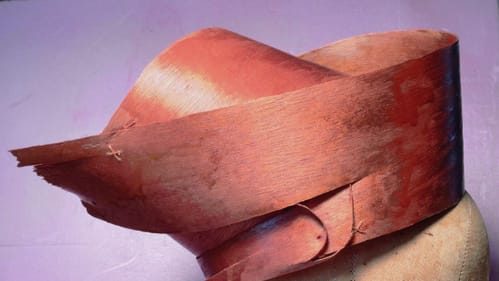 A fanciful, dynamic ladies’ hat on a cloth mannequin head is made out of shiny, bright curls of fragile wood veneer.