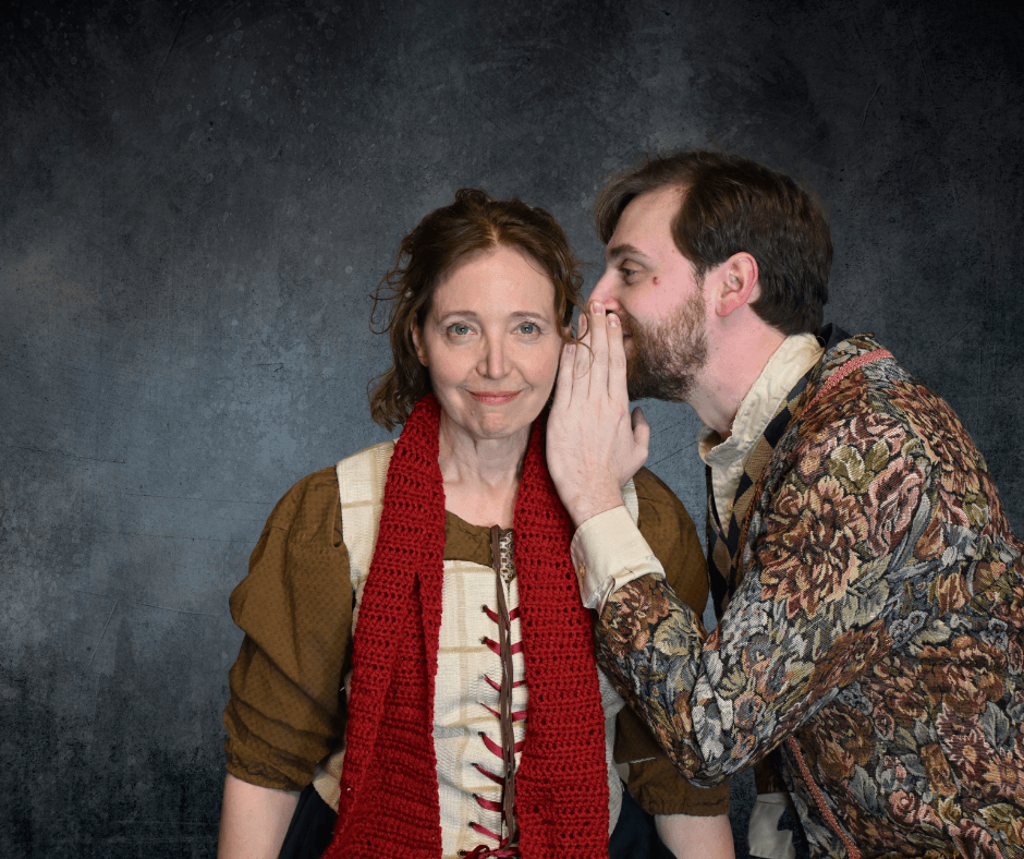 A woman looks towards camera, a man whispers in her ear. They both stand in front of a gray and black backdrop