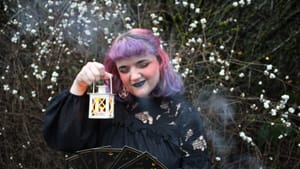 Markham, a white person with shoulder-length purple hair, holds a tiny lantern while looking at black tarot cards.
