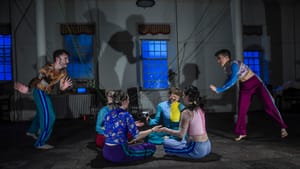 Four Wolfthicket dancers sit playing in a circle on the floor. On the sides, two standing dancers move with arms outstretched