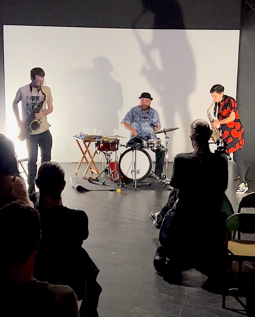 Three performers on stage—two on saxophone, one on drums—white muslin wall behind them, audience in front