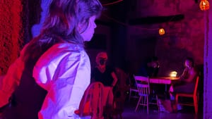 In a wash of red and purple light, Leiberman, back to the camera, heads toward audience members at a lantern-lit table