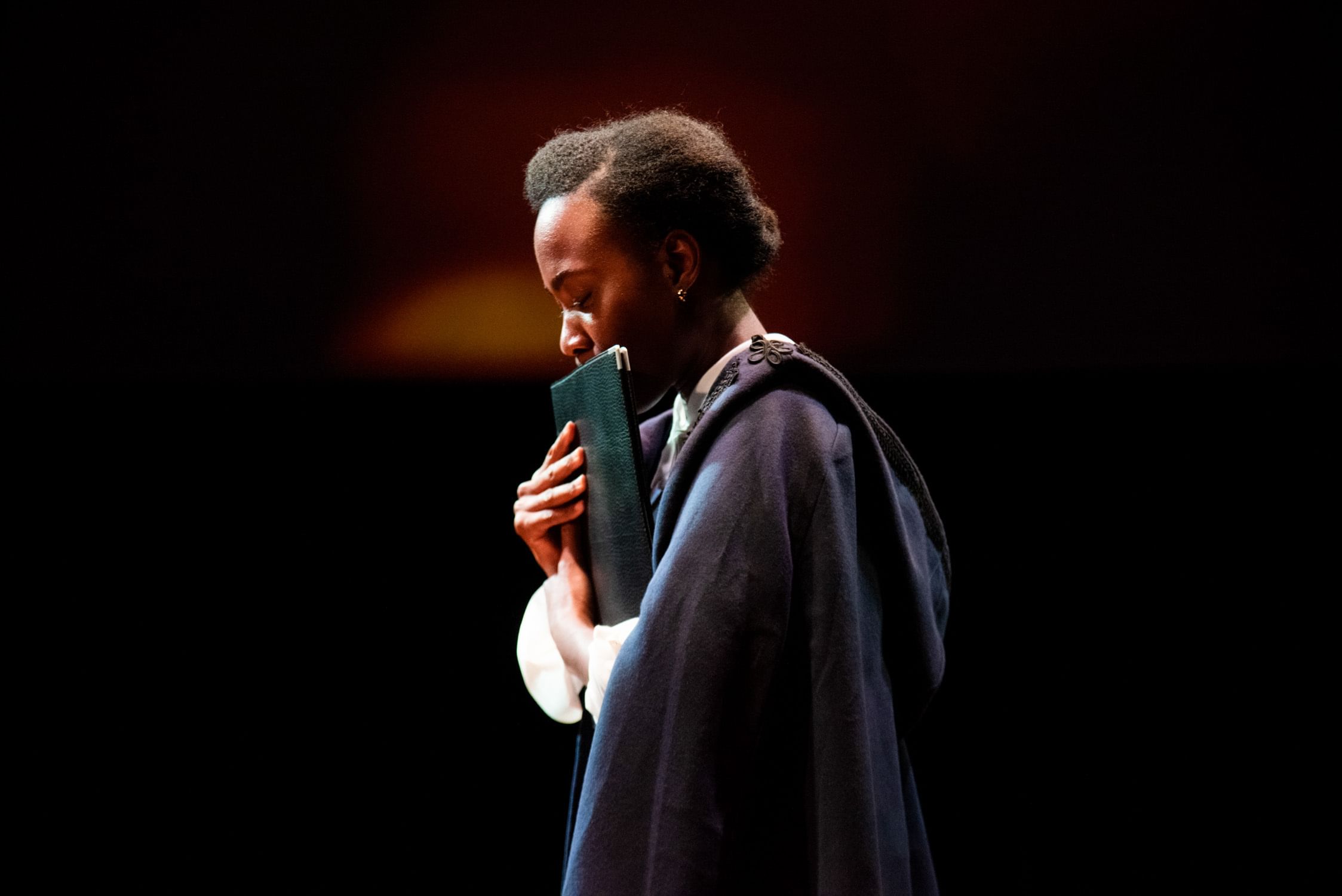 Rogers, a young Black woman seen in profile, wearing a dark-blue cape, presses a green folio contemplatively to her lips.