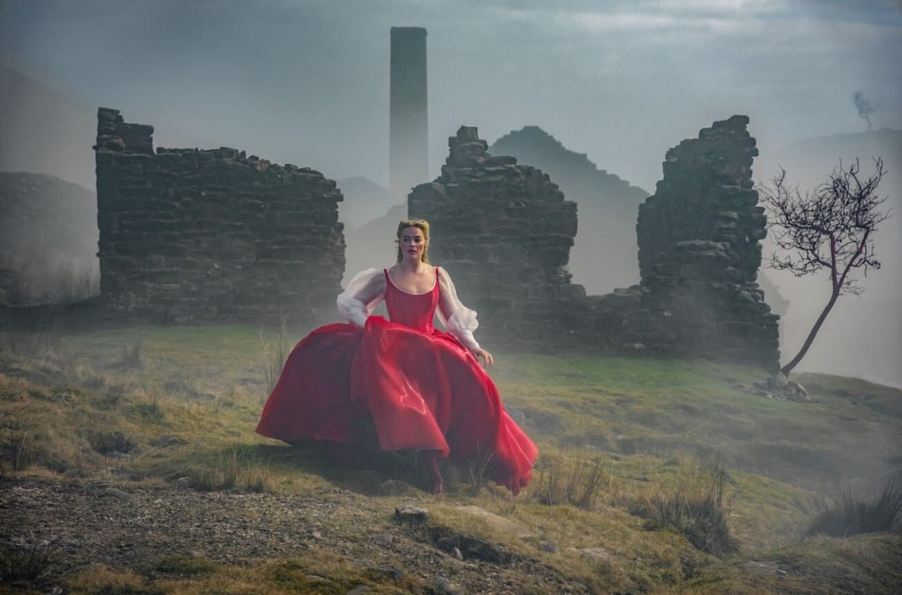 Robbie, in a billowing bright red dress with puffed white sleeves, walks in front of a misty stone ruin on the moor.