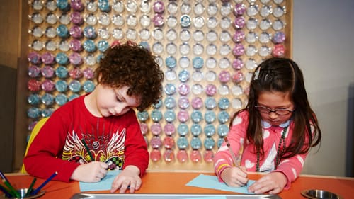 Two preschool aged children look down intently at the blue papers they are each drawing on.