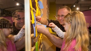 A smiling man helps a kindergarten-aged girl affix a little paper note to a wooden rack with many other notes.