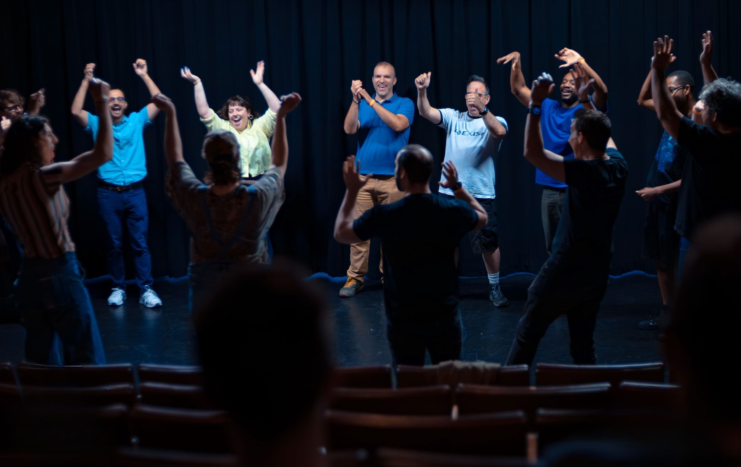 A group of performers on stage raise their arms or applaud, excited and laughing. Crowd before them also with their arms up