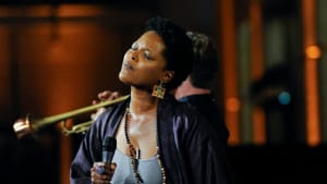 Wisher, a Black woman, sings on stage w/ a mic, her head tilted and eyes closed. Instruments, players are blurred behind her