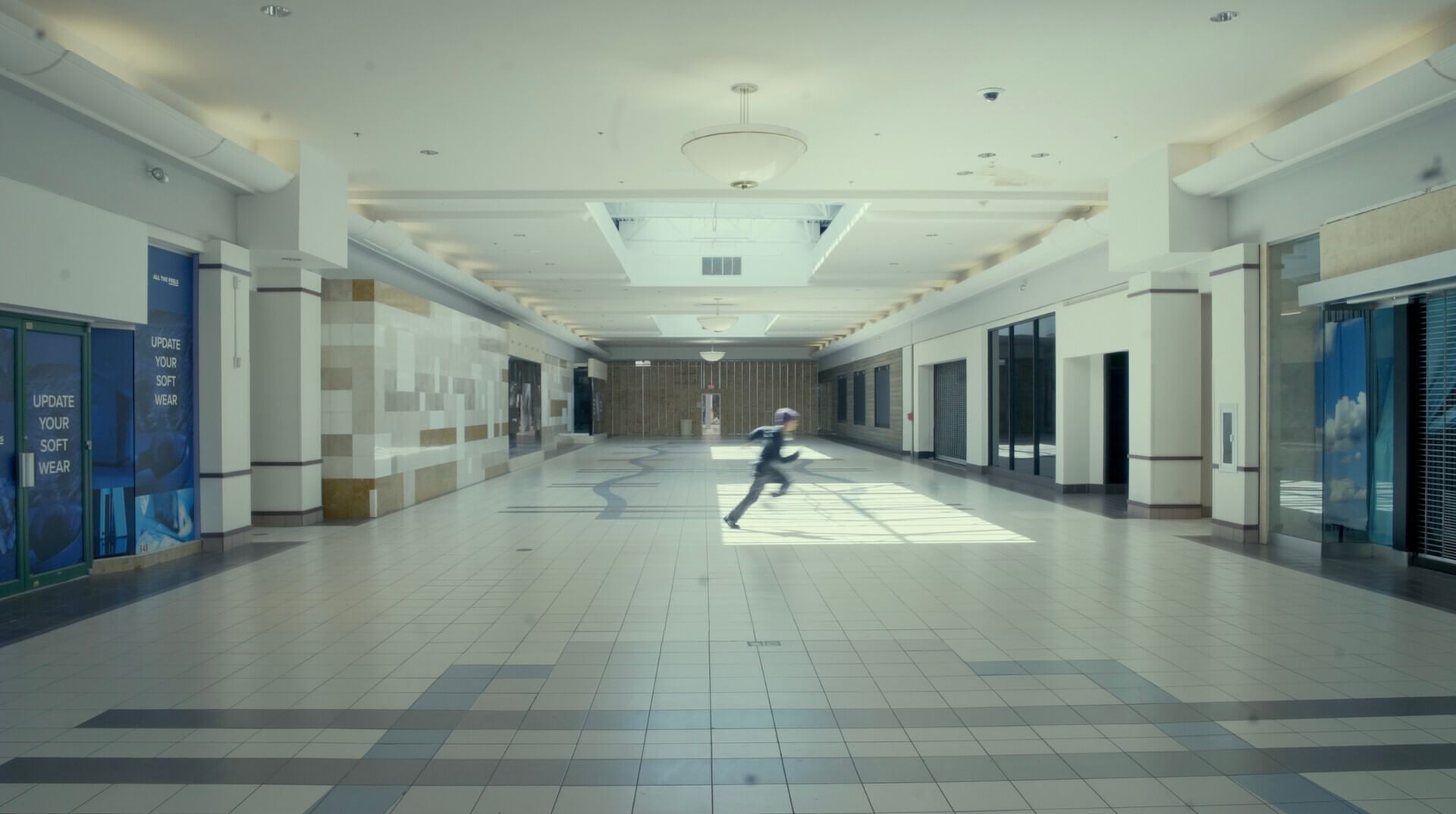 Movie still of a child, maybe preteen aged. Runs across hall in bright, empty mall, slightly blurred in the frame