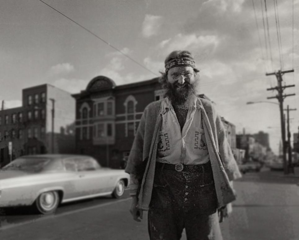 Vintage black & white photo of Zagar, in offbeat clothes, smiling impishly through his beard, on the 1970s city street.