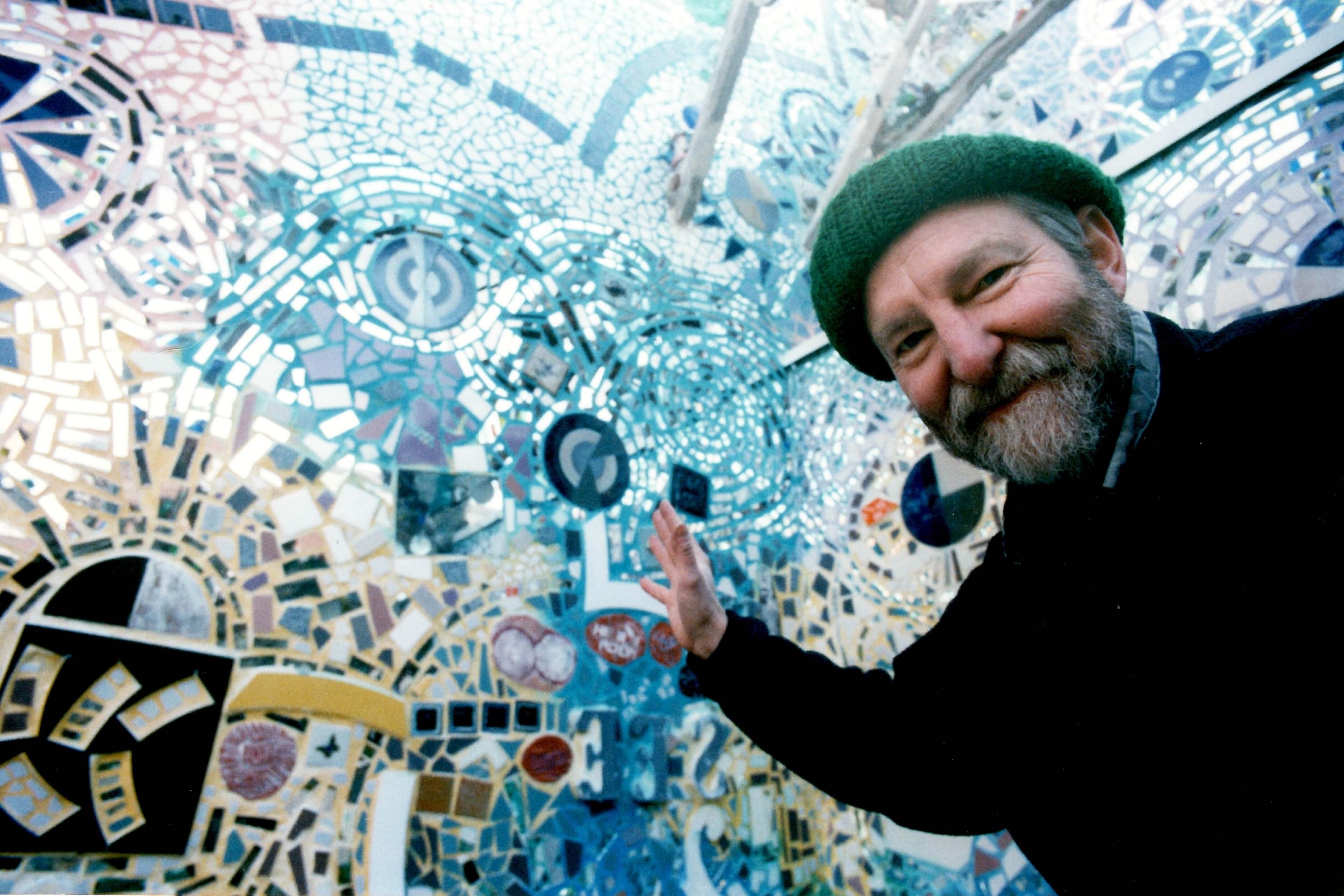 Zagar, a white man in a green knit hat, gestures to a the wild, shiny, swirling mosaic mural covering the walls behind him.