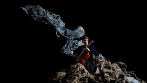 Against a black backdrop, a man wearing black plays the cello, in the middle of a large diagonal crinkled abstract sculpture.