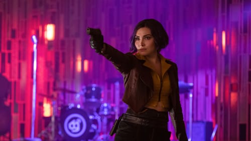 Valentine dressed in her iconic jacket w/ yellow blouse & black shorts, points a gun off camera, drum set on stage behind her