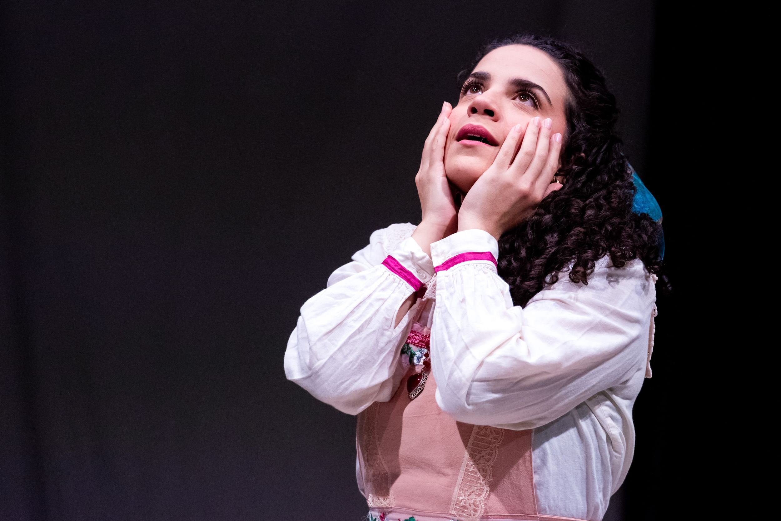Close-up on Cicchetti, a young woman with dark glossy curls, putting her hands to her face and looking up as she sings.