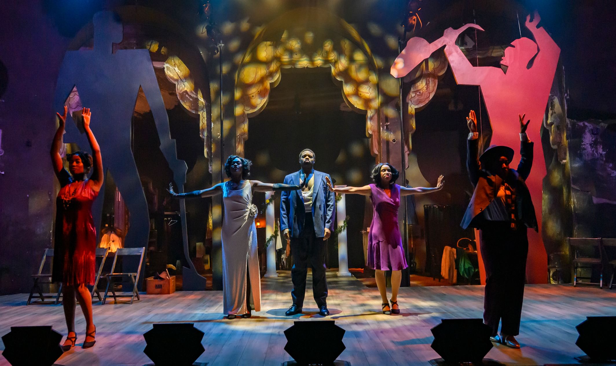 Five cast members in 1920s formal wear pose together, on an Art Deco stage with giant human silhouettes behind.
