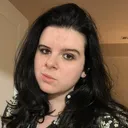 Headshot of Adryan Corcione, a white person with long dark wavy hair wearing a black & white floral top.