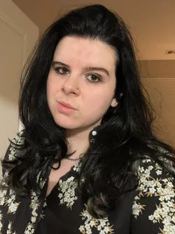 Headshot of Adryan Corcione, a white person with long dark wavy hair wearing a black & white floral top.