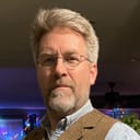 Bart is a white man with short white-gray hair and beard, wearing a light shirt and brown vest.