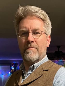 Bart is a white man with short white-gray hair and beard, wearing a light shirt and brown vest.