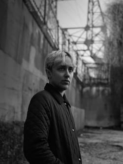 Black & white photo of Bruce, a man with blond hair wearing a black jacket, standing in an abandoned industrial setting.