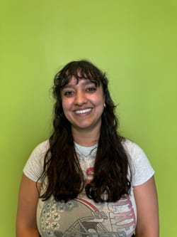 Photo of Chhaya, a smiling Indian woman with long dark hear, wearing a t-shirt, standing against a green background.