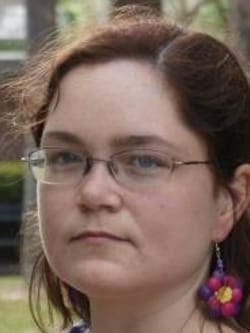 Headshot of Eileen Fay, a white woman with brown hair, glasses, and flower-shaped earrings. Her face is somber.